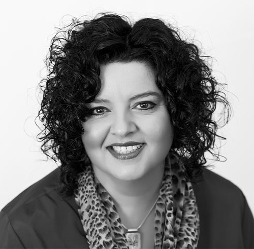 Black and white portrait of a woman with curly dark hair, smiling. She is wearing a patterned scarf and a pendant necklace, and looking directly at the camera.