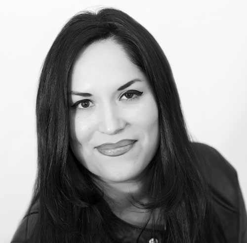 A woman with long dark hair and a slight smile, wearing a dark top, is photographed in black and white against a plain background.