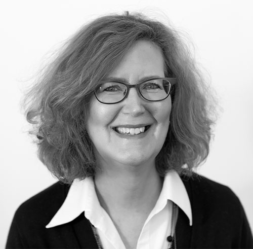 A woman with shoulder-length curly hair and glasses smiles at the camera. She is wearing a white collared shirt under a dark sweater. The background is plain and light-colored. The image is in black and white.