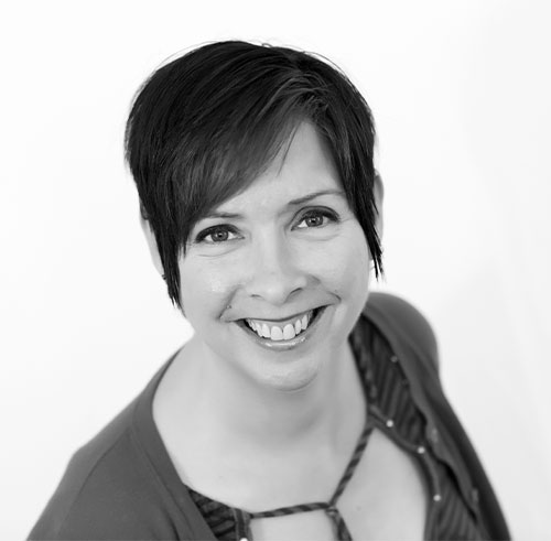 Black and white portrait of a smiling woman with short dark hair, wearing a striped top and a cardigan, photographed against a plain light background.