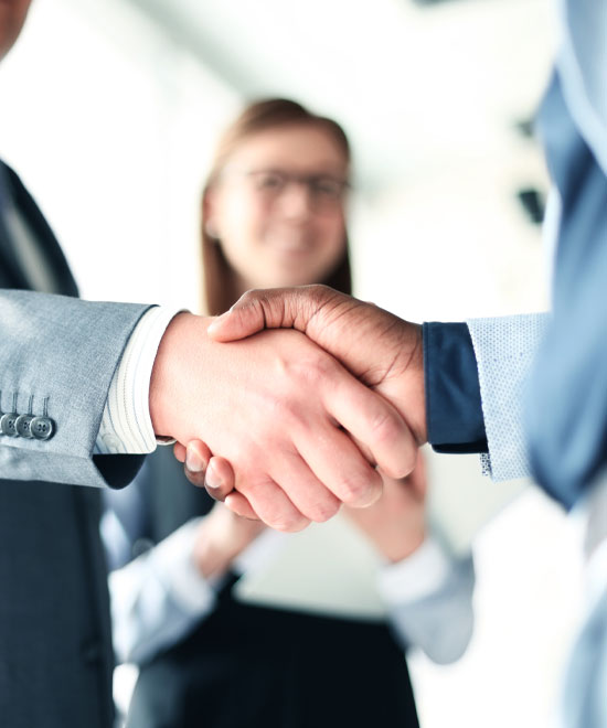 Two people in business attire shake hands, while a woman in the background smiles and observes their interaction in an office setting.