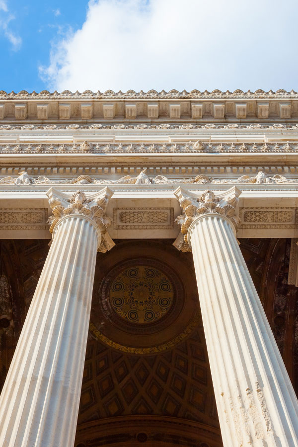 Looking up at the ornate columns and decorated ceiling of a classical building with intricate designs, against a partly cloudy blue sky.