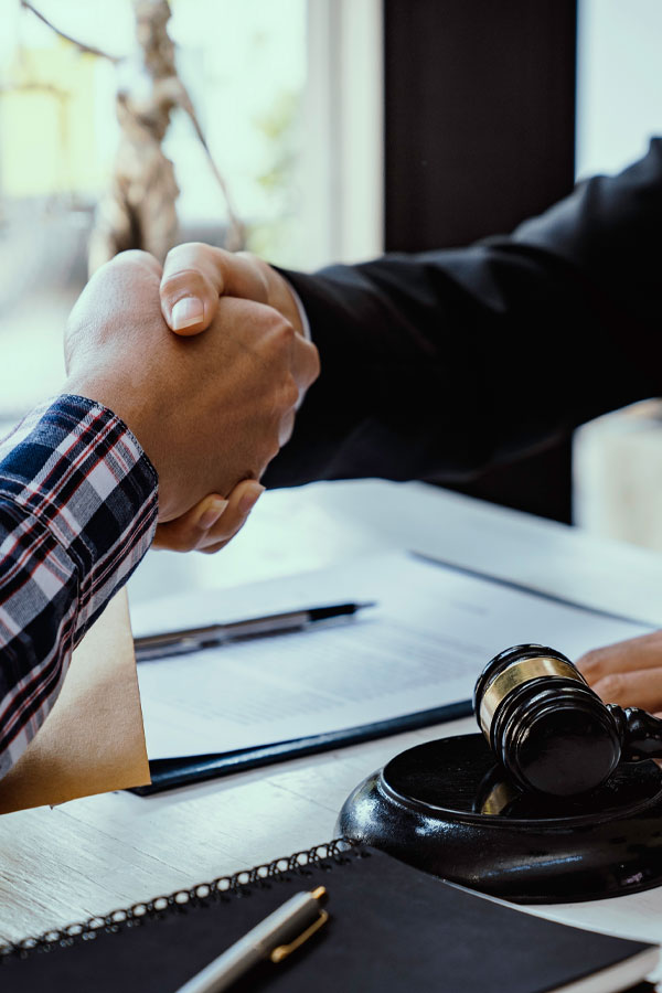 Two people shaking hands across a desk with legal documents, a pen, and a judge's gavel visible, suggesting a legal agreement or settlement.