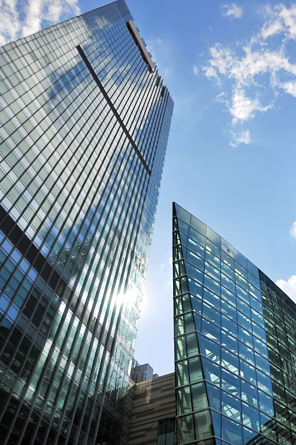 Two modern glass skyscrapers reflecting sunlight and clouds, photographed from a low angle against a bright blue sky.