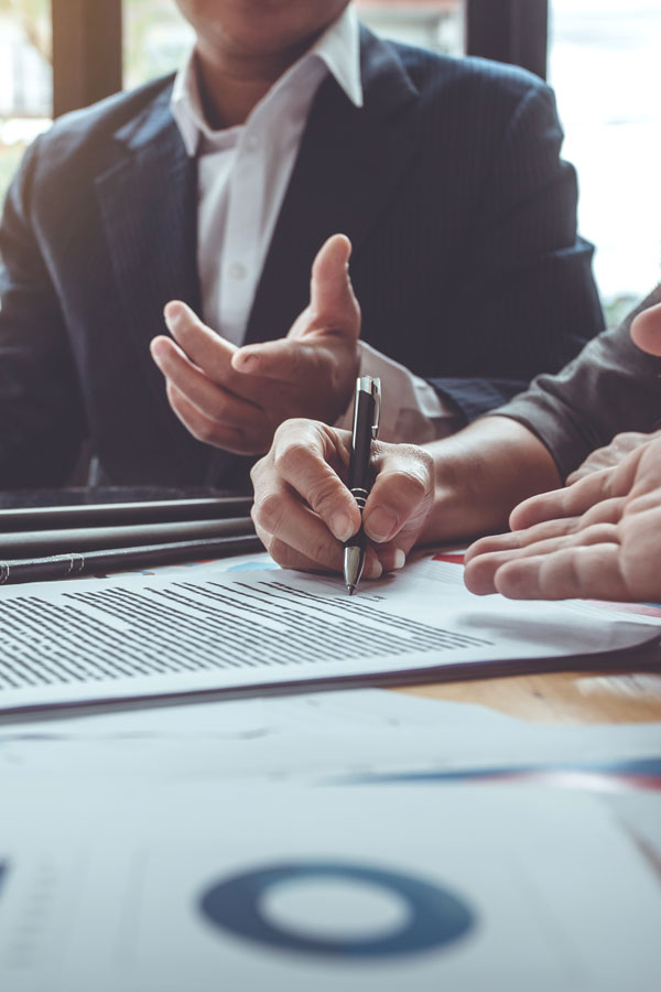 Two people in business attire sit at a table, one signing a document with a pen while the other gestures with open hands; charts and papers are visible in the foreground.