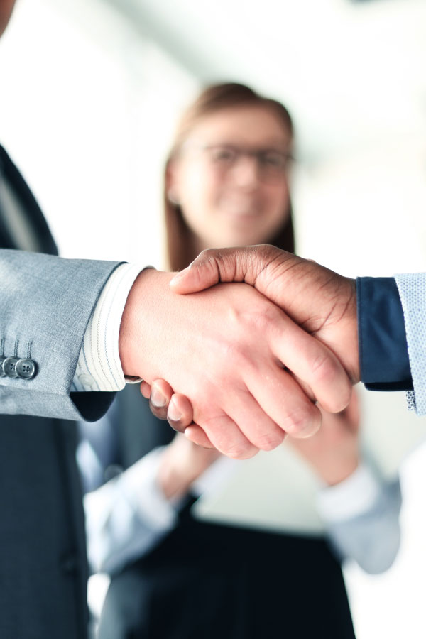 Close-up of two people shaking hands, wearing business attire, with a woman in the background smiling and watching them. The setting appears to be a bright office environment.