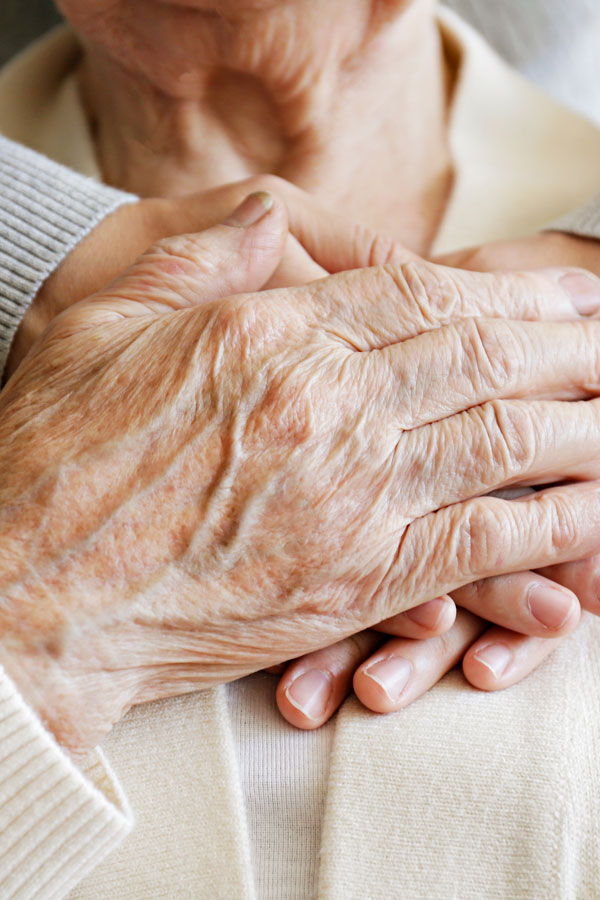 Close-up of an elderly person’s hands resting on their chest, with a younger person’s hand gently placed underneath, conveying care and support.