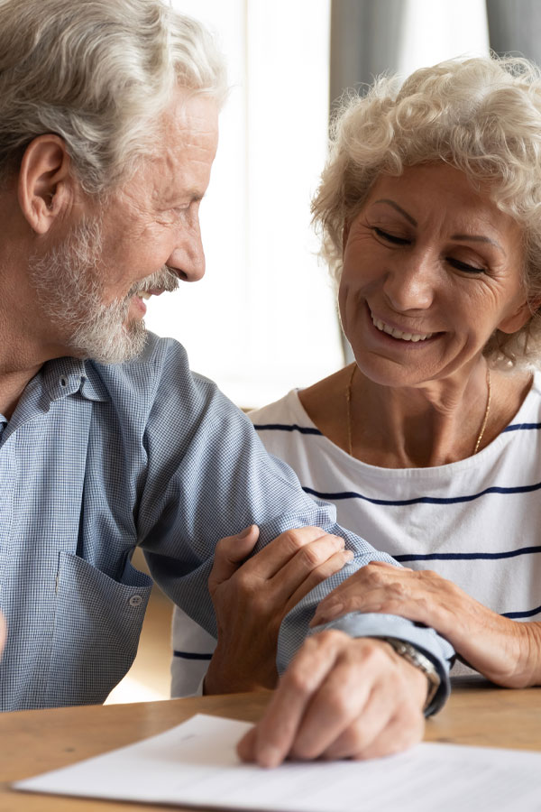 An older man and woman sit closely together, smiling warmly at each other. The woman holds the man's arm affectionately while he points to a document on the table in front of them.