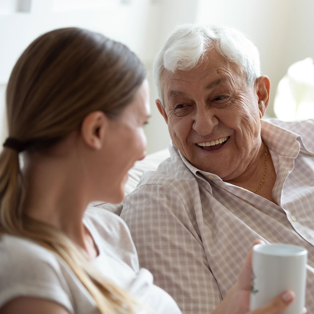 An older man with gray hair and a younger woman sit close together on a couch, smiling warmly at each other. The woman holds a white mug, and soft natural light fills the room.