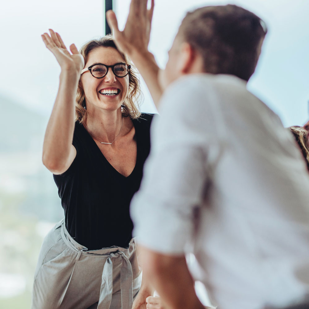 Two people smiling and giving each other a high five in a bright indoor setting, suggesting celebration or teamwork. The woman in front is wearing glasses, a black shirt, and light pants.