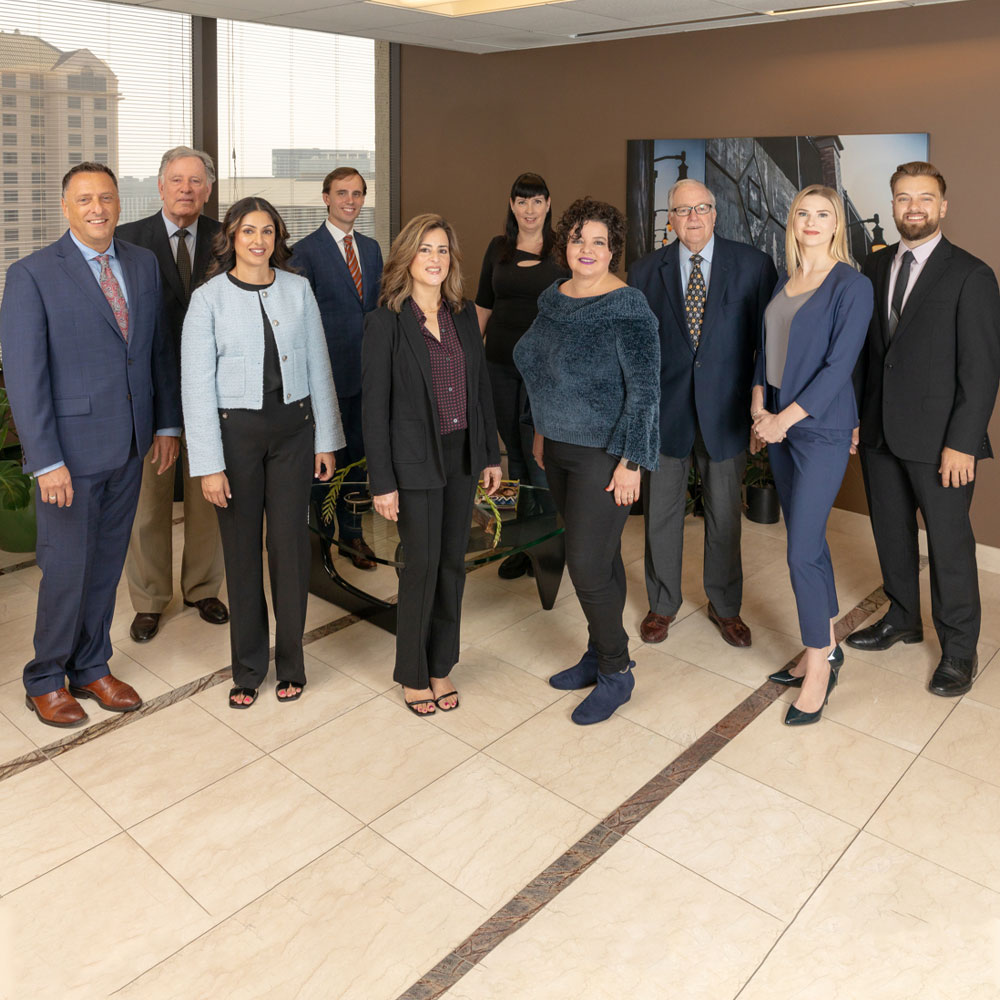 A group of ten professionally dressed adults, five men and five women, stand together in a modern office lobby with large windows and city views in the background.